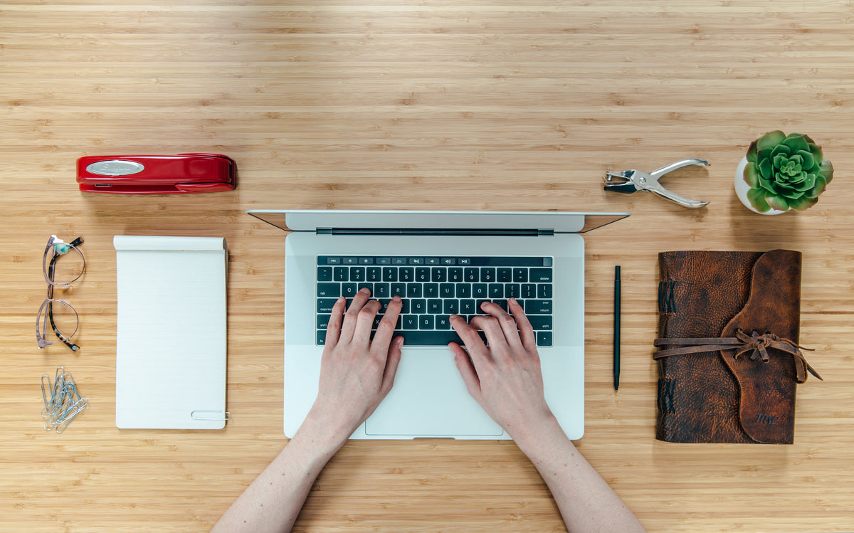 Hands typing on a laptop on a desk with a stapler, notebook and office supplies